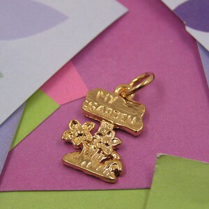 May include: Gold charm with a sign that says "My Garden" and two stylized flowers. The charm is on a pink and green background.