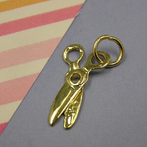 May include: A gold-colored metal charm in the shape of a pair of scissors. The charm has a ring at the top for attaching to a necklace or bracelet.