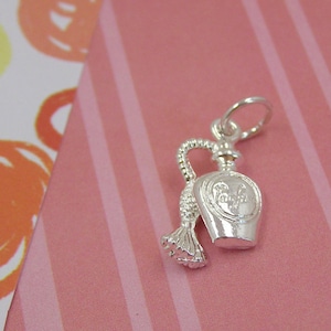 Puede incluir: Un charm de plata con forma de frasco de perfume con un borla. El frasco tiene un diseño detallado y un pequeño lazo para sujetarlo a una pulsera o collar.