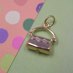 May include: Gold-toned charm in the shape of a small handbag with a clasp. The charm has a detailed design and a loop for attaching to a bracelet or necklace.