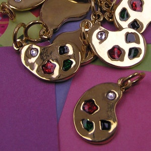 May include: Gold-tone charm in the shape of a paint palette with red, green, and black enamel accents. The charm has a small clear rhinestone on the top.