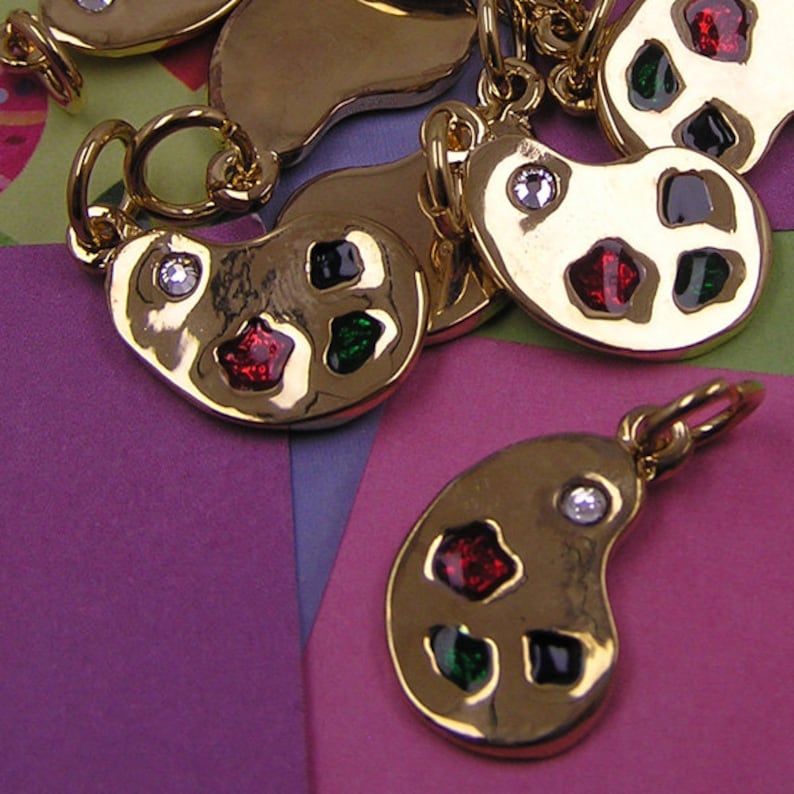 May include: Gold-tone charm in the shape of a paint palette with red, green, and black enamel accents. The charm has a small clear rhinestone on the top.
