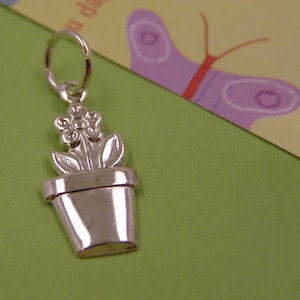 May include: A silver charm in the shape of a potted flower. The flower has five petals and a center with a small circle. The pot is round and has a smooth surface.