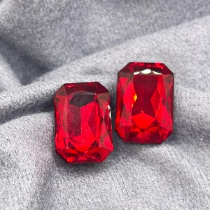 May include: Two rectangular, faceted red gemstones with a brilliant cut. The stones feature sharp, clean edges and a deep, vibrant crimson colour that reflects light across their multiple surfaces, creating a clear and polished appearance.