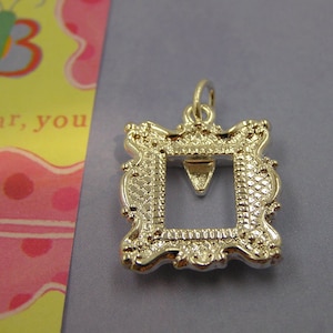 Puede incluir: Encanto de plata en forma de marco de imagen con un diseño decorativo. El marco está abierto y listo para contener una foto u otro artículo pequeño.