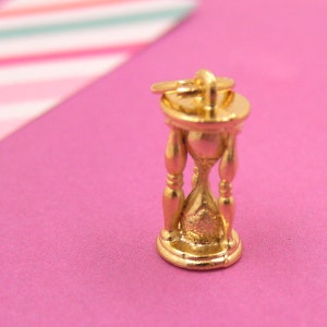 Puede incluir: Un colgante de reloj de arena de color dorado. El reloj de arena está detallado con una superficie texturizada y un pequeño bucle en la parte superior para sujetarlo a una cadena.