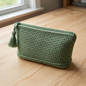 Puede incluir: Un estuche rectangular de ganchillo en color verde salvia con un patrón de puntos texturizados, una cremallera a juego en el borde superior y una borla decorativa de hilo unida al tirador, un accesorio de almacenamiento sencillo.