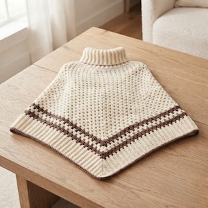 May include: A cream-colored crochet poncho featuring a ribbed turtleneck collar and a diamond-shaped pattern. The garment is accented with a dark brown horizontal stripe near the bottom edge, which is finished with a ribbed knit border.