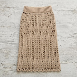 May include: A beige crochet skirt featuring a repeating openwork pattern throughout the body. The garment has a wide, ribbed elasticized waistband and a scalloped hemline. The intricate needlework creates a textured, semi-sheer appearance.
