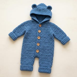 Op de afbeelding: Een blauwe gehaakte jumpsuit uit één stuk met een capuchon met twee kleine ronde oortjes aan de bovenkant. Het kledingstuk heeft lange mouwen met geribbelde boorden, geribbelde broekspijpen en een verticale sluiting aan de voorzijde met vijf ronde houten knopen.