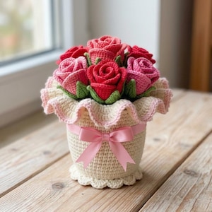 Puede incluir: Un ramo de rosas rojas y rosas tejidas a ganchillo con hojas verdes, dentro de una maceta tejida de color crema. La maceta presenta un borde superior con volantes, una base festoneada y un lazo de satén rosa claro atado en el centro.