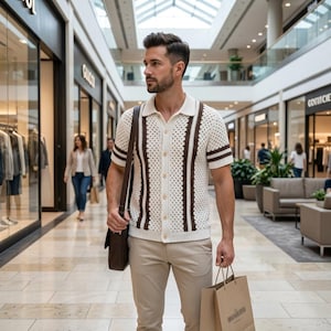 May include: A short-sleeved, cream-colored knit polo shirt with vertical brown stripes, a collared neckline, and a button-down front. The shirt is paired with light beige trousers and a dark brown leather shoulder bag carried on the side.