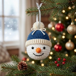 May include: A crocheted snowman ornament featuring a white spherical head, a dark blue beanie with a cream-colored brim, a small orange carrot-shaped nose, black bead eyes, and a stitched smile, suspended by a silver ribbon from a pine branch.