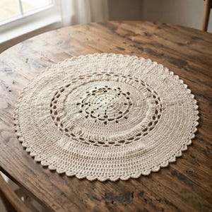 May include: A round, off-white crochet doily featuring a central circular pattern with intricate openwork stitching, surrounded by a solid textured band and finished with a delicate scalloped edge, creating a detailed circular textile design.