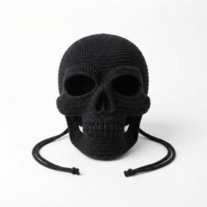 Puede incluir: Una máscara de calavera tejida a ganchillo en color negro, con cuencas oculares profundas y huecas, una cavidad nasal definida y una mandíbula texturizada. La máscara está hecha de hilo oscuro e incluye dos cordones negros largos y finos.