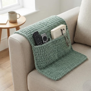 May include: A sage green crochet sofa armrest organizer featuring multiple pockets. The organizer is draped over the arm of a beige couch and contains two black remote controls, a white book, and a pair of black-rimmed reading glasses.