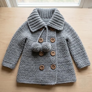 Puede incluir: Un abrigo de ganchillo gris con cuello ancho, mangas largas y cierre frontal de doble botonadura con seis botones redondos de madera. Dos pompones de lana gris están sujetos al cuello mediante finos cordones de ganchillo, añadiendo textura.