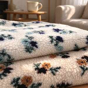 May include: Two folded sherpa blankets with a thick, textured cream-colored fleece surface. The top blanket features a floral pattern in shades of navy blue and light blue, while the bottom blanket displays a floral print in brown and dark green.