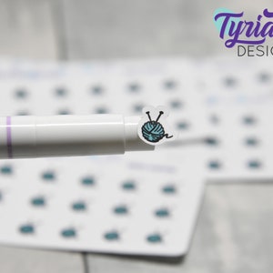 K&ouml;nnte beinhalten: Eine Bogen mit Aufklebern mit einem Design aus einer blauen Wollkugel und Stricknadeln. Die Aufkleber sind auf wei&szlig;em Hintergrund und werden von einem wei&szlig;en Stift mit einem violetten Streifen gehalten.