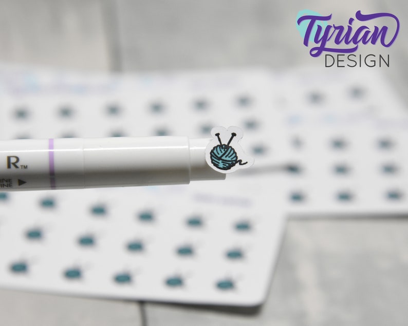 K&ouml;nnte beinhalten: Eine Bogen mit Aufklebern mit einem Design aus einer blauen Wollkugel und Stricknadeln. Die Aufkleber sind auf wei&szlig;em Hintergrund und werden von einem wei&szlig;en Stift mit einem violetten Streifen gehalten.