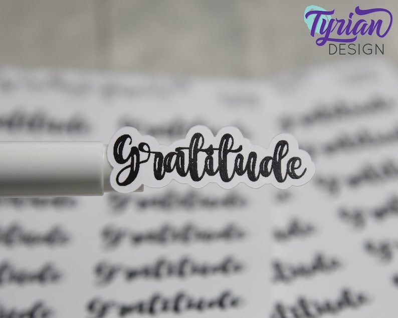 May include: A sheet of white paper with the word "Gratitude" written in black cursive script. The word is printed on a sticker and is being held by a white pen.