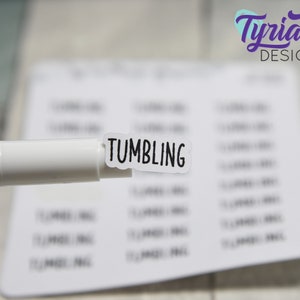 May include: A white sticker with the word "TUMBLING" printed in black lettering. The sticker is being held by a white marker with a purple cap.