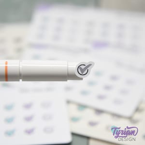 Könnte beinhalten: Ein weißer Stift mit einem orangefarbenen Streifen und einem kleinen Aufkleber mit einem grauen Häkchen in einem schwarzen Kreis. Der Aufkleber befindet sich auf der Spitze des Stifts. Im Hintergrund befinden sich mehrere weiße Karten mit teal- und lilafarbenen Designs.