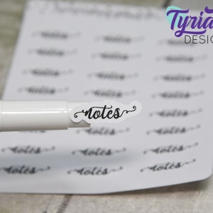 May include: A white marker pen with a clear sticker on the end that says "notes" in a cursive font. The sticker is on a sheet of white paper with multiple stickers that say "notes" in a cursive font.