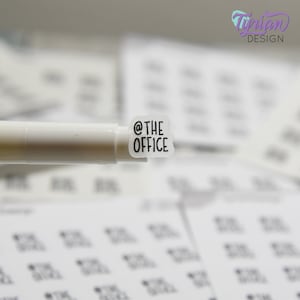 May include: A white sticker with the text "@THE OFFICE" in black lettering. The sticker is being held by a white pen.
