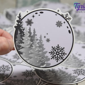 May include: A round sticker featuring a winter scene with a black and white color scheme. The design includes stylized evergreen trees, snowflakes, and a moon. The sticker has a white border and the text "Tyrian Design".