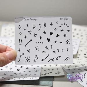 May include: A white sticker sheet with various black doodle designs, including hearts, stars, arrows, and bursts. The text "TyrianDesign" and "TINY-10561" are printed on the sticker sheet. The sticker sheet is held by a hand.