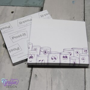 May include: A white Post-it note pad with a row of cartoon marshmallows drawn on the bottom edge. The marshmallows have purple eyes and mouths.