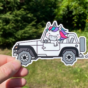 Puede incluir: Un Jeep blanco con un unicornio conduciendo. El unicornio tiene una crin y un cuerno rosa, azul y blanco. El Jeep est&aacute; conduciendo por un camino de tierra y tiene un parachoques gris y una rueda de repuesto gris.