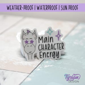 Könnte beinhalten: Weißer Aufkleber mit einem Cartoon-Einhorn-Design und dem Text "Main Character Energy". Das Einhorn hat lila Sternaugen und ein lila Horn. Der Aufkleber ist wetterfest, wasserdicht und sonnenbeständig.