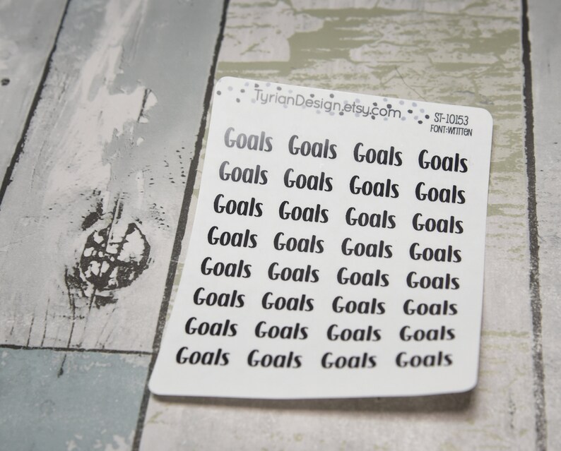 May include: A white sticker sheet with the word "Goals" printed in a black handwritten font. The sticker sheet has the text "TyrianDesign.etsy.com" and "ST-10153 FONT-WITTEN" printed on it. The sticker sheet is on a light gray and white wooden surface.