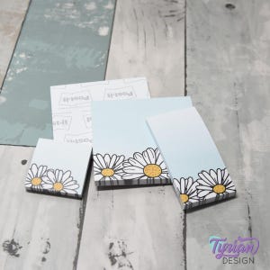 May include: Several Post-it note pads in various sizes. The pads have a light blue gradient background with a daisy design along the bottom edge. The daisies have white petals and yellow centers.
