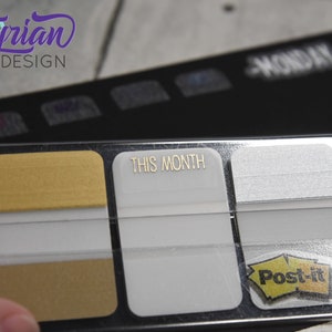K&ouml;nnte beinhalten: Ein transparenter Plastikhalter mit drei Haftnotizen. Die obere Notiz ist golden, die mittlere Notiz ist wei&szlig; mit dem Text "THIS MONTH" in goldenen Buchstaben, und die untere Notiz ist wei&szlig; mit dem Logo der Marke Post-it.