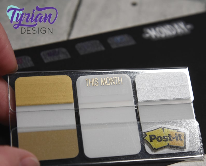 K&ouml;nnte beinhalten: Ein transparenter Plastikhalter mit drei Haftnotizen. Die obere Notiz ist golden, die mittlere Notiz ist wei&szlig; mit dem Text "THIS MONTH" in goldenen Buchstaben, und die untere Notiz ist wei&szlig; mit dem Logo der Marke Post-it.