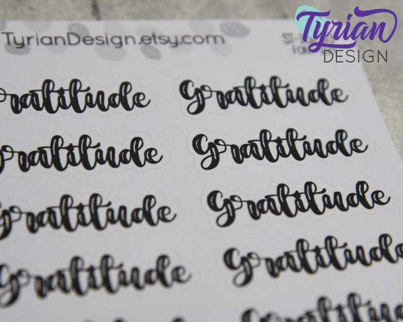 May include: A sheet of stickers with the word "Gratitude" written in black cursive lettering. The stickers are on a white background.