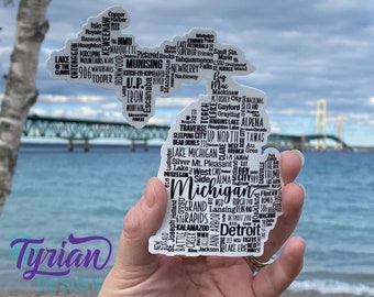May include: A white sticker shaped like the state of Michigan with black text outlining cities, towns, and landmarks. The sticker is held in a hand against a backdrop of a blue lake and a bridge.