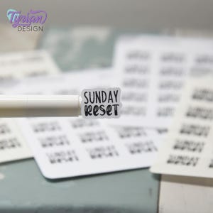 Peut inclure: Un petit autocollant rectangulaire avec les mots "SUNDAY RESET" en noir, dans une police manuscrite. L'autocollant est sur un fond blanc et est tenu par un stylo blanc. D'autres autocollants similaires sont en arrière-plan.