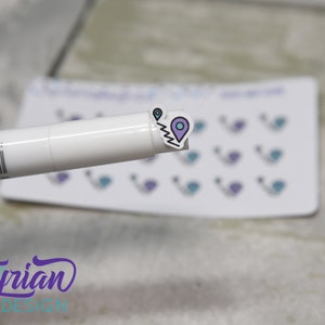May include: A sheet of stickers with a purple and blue location pin design. The stickers are on a white background. A white marker is in the foreground.