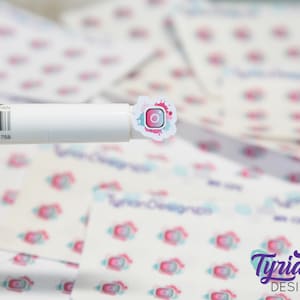 May include: A close-up of a white sticker with a pink, blue, and white watercolor design. The sticker features a stylized Instagram logo with a white circle inside a pink square.