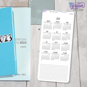 Può includere: Un adesivo calendario 2027, bianco con testo nero, che mostra i mesi da gennaio a dicembre. Un'agenda blu con il testo "023 ONICHI HO 2023 cousin" è visibile sullo sfondo. Ideale per l'organizzazione.