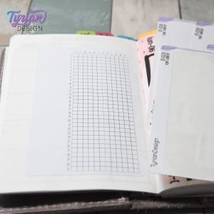 Puede incluir: Una agenda abierta que muestra una hoja de pegatinas blanca con una cuadrícula negra y filas numeradas del 1 al 31. La hoja incluye el texto TyrianDesign y SF-10036. A la derecha se ven otras hojas de pegatinas coloridas con pestañas.