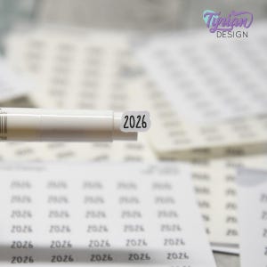Puede incluir: Primer plano de un bolígrafo blanco con una pequeña pegatina con la inscripción "2026". La pegatina tiene texto negro sobre fondo blanco. El fondo incluye hojas de pegatinas con el número "2026" impreso.