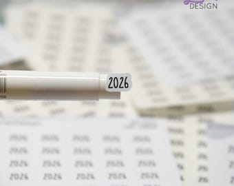 May include: A close-up of a white pen with a small sticker labeled "2026" attached to the tip. The sticker is black text on a white background. The background includes sheets of stickers with the number "2026" printed on them.