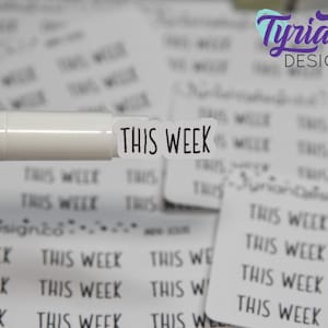 Pode incluir: Um close-up de um adesivo branco com o texto "THIS WEEK" impresso em preto. O adesivo está em um fundo branco com outros adesivos que dizem "THIS WEEK" no fundo.