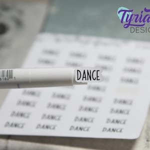 May include: A white marker pen with a black and white sticker that says "DANCE" on it. The sticker is being held over a sheet of paper with the word "DANCE" printed on it multiple times.