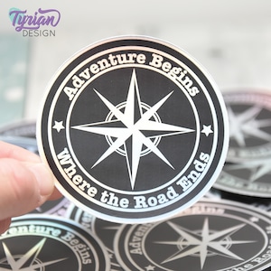 May include: A black and white compass sticker with the text "Adventure Begins Where the Road Ends". The sticker is iridescent and has a silver border.